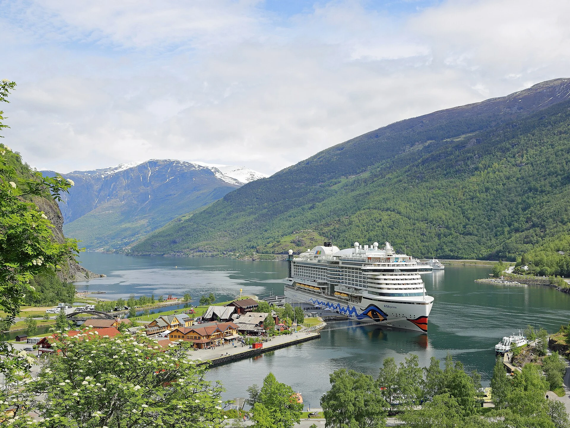 Kreuzfahrtschiff am Kai von Flåm am Ende des Aurlandsfjords, dahinter Ortszentrum und bewaldete Fjordberge.
