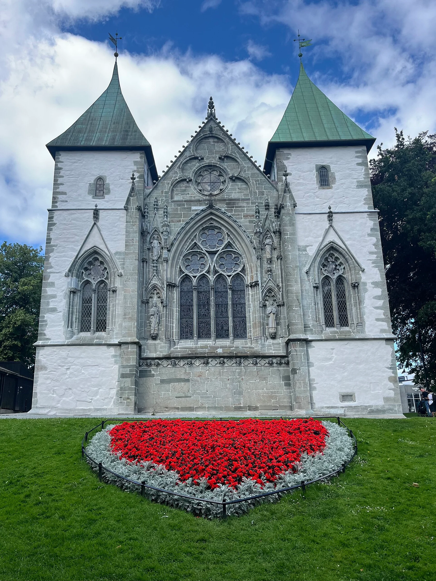 Der gotische Chor der Stavanger domkirke mit hellem Verputz und zwei grün gedeckten Türmen, davor ein rotes Blumenbeet.